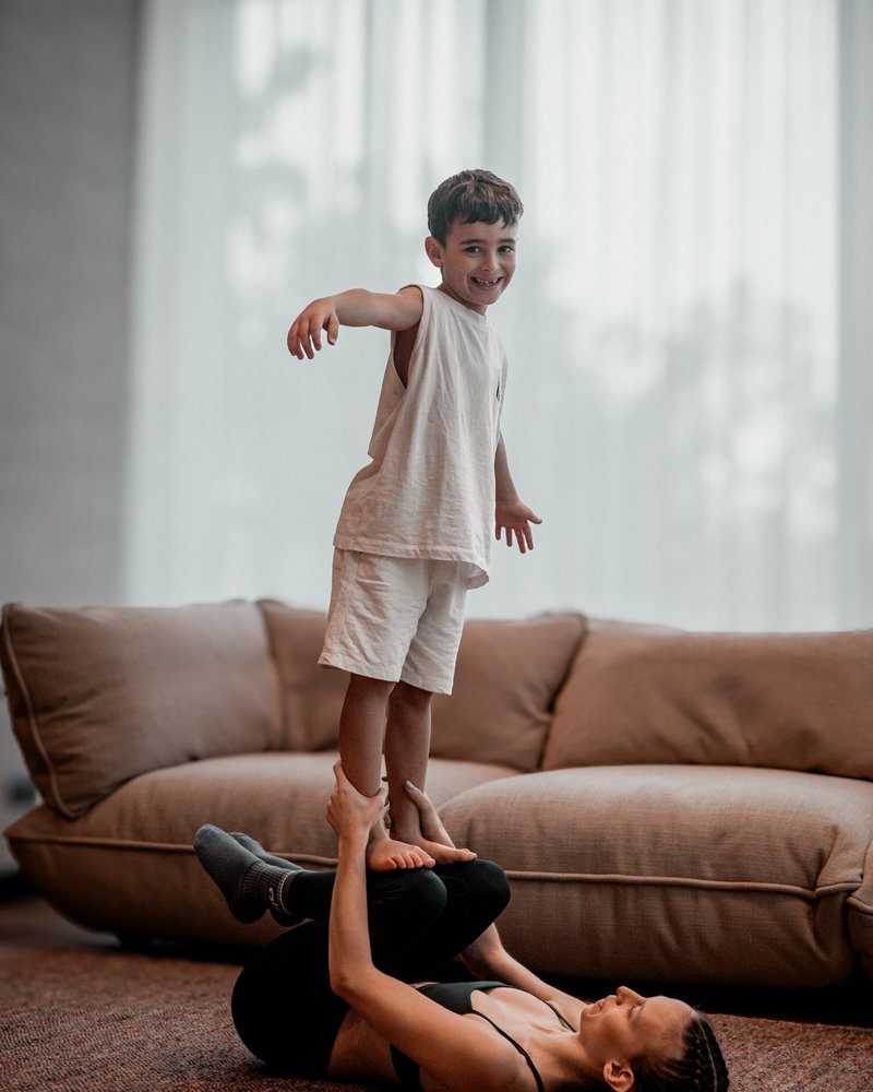 A mother lies on her back and bases her young son standing upright on her feet, both smiling.